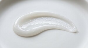 A macro photograph of a fresh gel-cream skincare texture. A smooth, elegant curved swatch of a lightweight, semi-translucent white gel-cream with a subtle pearlescent sheen, resting on a clean, matte white porcelain surface. The lighting is soft and directional, catching the moist, hydrating micro-reflets of the cream. Extreme close-up, shallow depth of field, 100mm macro lens, f/8, ultra-sharp details showing the delicate consistency of the formula. No packaging. no AI artifacts, no warped or smeared text, no fake words, no invented logos, no plastic/waxy look, no doll-like skin, no oversaturated HDR, no stock-photo feel, no watermark, no extra fingers, no melted geometry