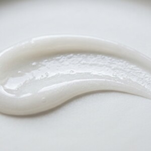 A macro photograph of a fresh gel-cream skincare texture. A smooth, elegant curved swatch of a lightweight, semi-translucent white gel-cream with a subtle pearlescent sheen, resting on a clean, matte white porcelain surface. The lighting is soft and directional, catching the moist, hydrating micro-reflets of the cream. Extreme close-up, shallow depth of field, 100mm macro lens, f/8, ultra-sharp details showing the delicate consistency of the formula. No packaging. no AI artifacts, no warped or smeared text, no fake words, no invented logos, no plastic/waxy look, no doll-like skin, no oversaturated HDR, no stock-photo feel, no watermark, no extra fingers, no melted geometry